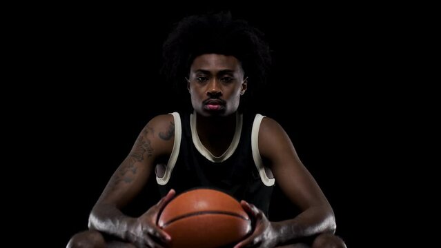Basketball player sitting on a bench holding a ball. Concentrated african american sports man against black background
