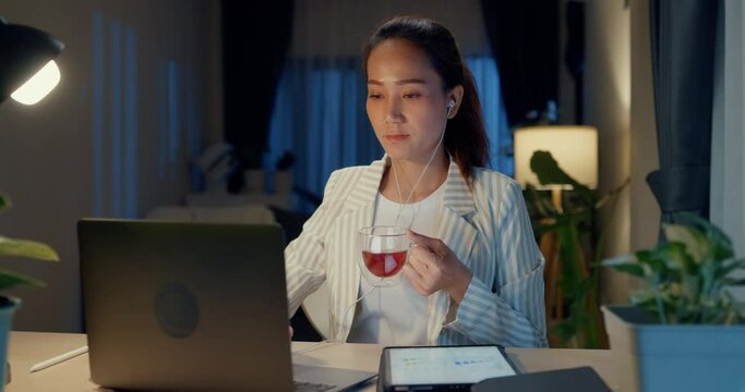 Young Asia businesswoman wear formal sit front of desk with laptop use earphone video online meeting drink cup of tea present finance report in living room at home night. Overtime work concept.