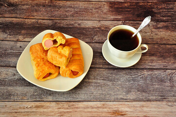 Several sausages fried in puff pastry and a cup of hot black coffee on a wooden table.