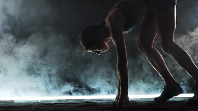 Silhouette female runner in race  start position. Girl in sportswear posing on lit track with smoke in the background.