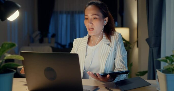 Young Asia businesswoman wear formal sit front of desk with laptop computer use earphone video online meeting hold tablet present finance report in living room at home night. Overtime work concept.