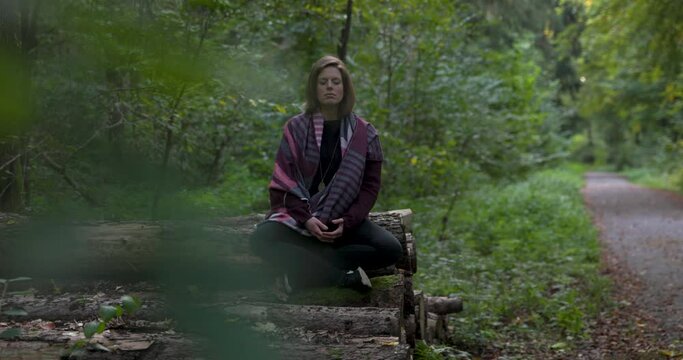 thoughtful natural women meditating in the green forest while sitting on logged trees