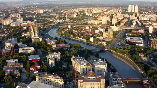 slow flowing Vardar River with a low water level through the center of Skopje under the many historic bridges as the skyline is illuminated by the setting sun. Drone panning dolley shot