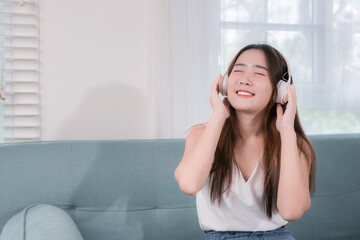 Asian woman use smartphone and wearing headphone listening music entertainment her enjoy and fun sitting on sofa in living room at home