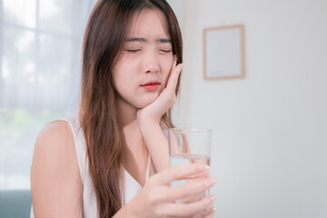 Asian woman has sensitive teeth after drinking cold water on sofa in living room at home. concept dental health