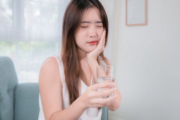 Asian woman has sensitive teeth after drinking cold water on sofa in living room at home. concept dental health