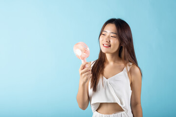 Asian beauty young woman holding pink portable electric mini fan near her face studio shot isolated on blue background, Female hand hold small plastic fan handheld she enjoying cool wind blowing - obrazy, fototapety, plakaty