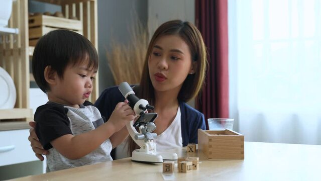 Cute little Asian boy playing microscope toys with his mother sitting beside in the living room.