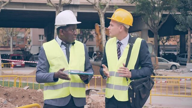 Two engineers, Caucasian and African-american men in hardhats and warnvests, standing and talking about road paving works documentation. 