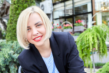 Blonde woman dressed in business style sits on a chair and looks at the camera. The woman has a break in the middle of the working day