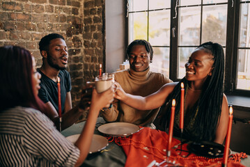 Best friends celebrating new year. Young people with candles, sitting at dining table. Diverse students during christmas party at home, smiling and laughing.