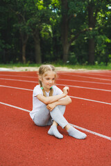 A little girl of 7-8 years old in a white t-shirt runs outdoors in the stadium. The girl is doing sports on a sunny day