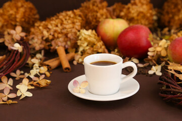 White coffee cup, autumn leaves, cinnamon, apples and dry hydrangea on brown background, front view. Selective focus