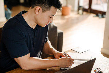 Adult asian man taking notes while studying/ working at home