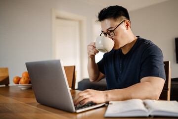 Adult asian man drinking tea/coffee while working with laptop