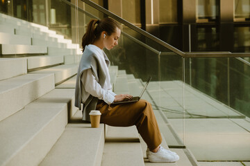 Young woman in earphones working with laptop while sitting on stair