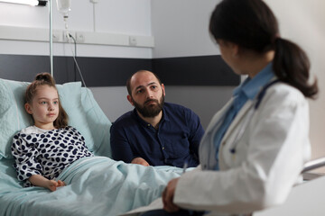 General practitioner consulting unwell recovering kid sitting in patient bed. Pediatric expert talking with father of hospitalized sick little girl resting in children healthcare facility ward.