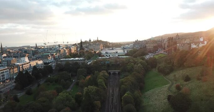 Beautiful sunrise drone shot of Princes Street Gardens, Edinburgh, approaching the train station with the castle and old town to the right.