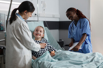 Happy physician expert and nurse consulting ill little girl health condition while smiling together inside hospital pediatric ward. Multiethnic clinic staff examining sick kid resting in patient bed.