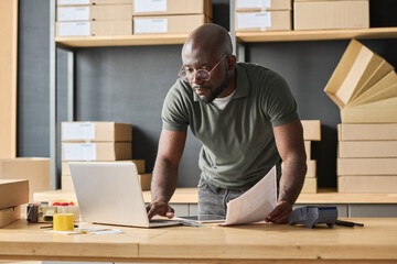 Serious African man concentrating on his work on laptop at table, he typing data of parcels during...