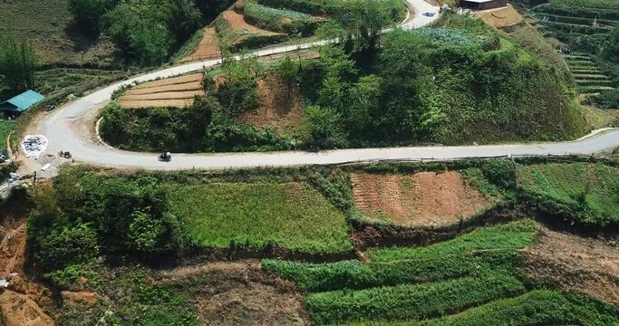 An aerial shot follows a motorbike up a winding road through terraced fields towards the top of a hill. South East Asia.