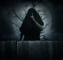 Grim reaper showing silence sign with finger standing over dead tree, full moon and cloudy spooky...