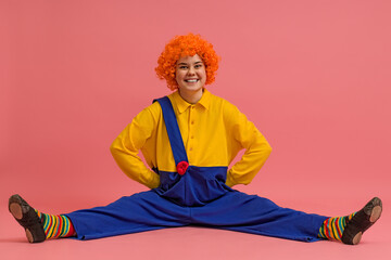 a joyful clown in a wig and a yellow-blue suit sits with his legs apart on a colored background
