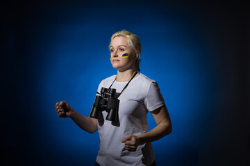 A blonde woman with a blue-yellow Ukrainian flag on her cheek with binoculars around her neck is walking. Blue background.