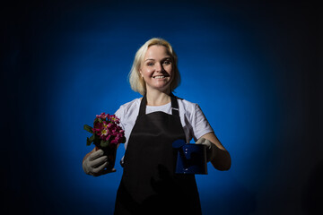 A blond gardener woman in a black apron and protective gloves holds a flower pot and a watering can in her hands. Dark background.