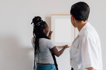 Woman looking at girlfriend hanging picture frame on wall at home