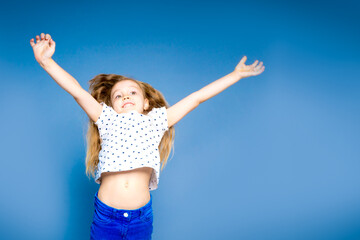 Cheerful little girl jumping lifts hands up. On a blue background in the studio