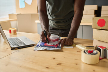 Fototapeta na wymiar Close-up of African man making notes on packet with order at table while distributing parcels to addresses