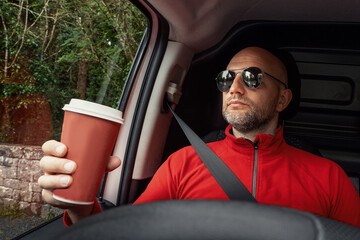 Male bald van driver with a cup of coffee. Selective focus. Break for a drink. Transportation and delivery service. Man in red shirt with beard and dark glasses.