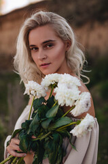 Fototapeta premium A stylish blonde girl with tanned skin and curly hair stands in a beige summer suit with a white flower on an overgrown quarry. Fashion, travel and lifestyle concept. Close up. 