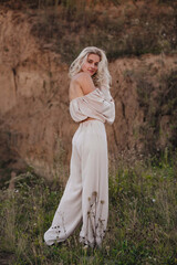 Fototapeta premium A stylish blonde girl with tanned skin and curly hair stands in a beige suit with a white flower on an overgrown quarry. Fashion, travel and lifestyle concept.