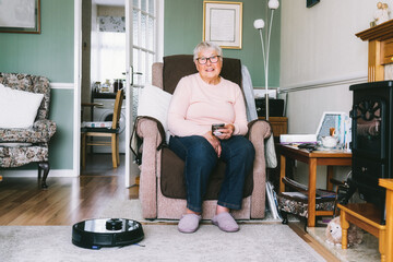 Senior woman using a robot vacuum cleaner through phone settings while sitting on armchair at home. Mature older lady holding mobile phone and control cleaning. Smart home. Seniors and technology