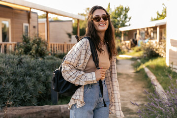 Positive young caucasian woman enjoys spending time outdoors during day. Brunette wears sunglasses, shirt, jeans and backpack. Good mood, fashion trends