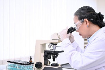 Professional senior female scientist looking under Microscope, analyzing samples in the lab.