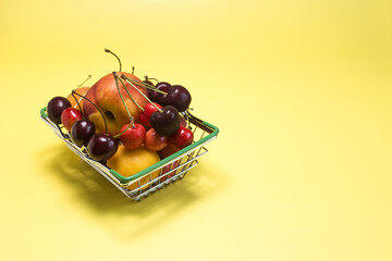 A grocery basket filled with various fruits - bananas, cherries, nectarines, apricots, peaches on a bright yellow background kopi space.