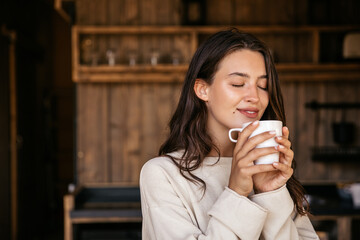 Cute young caucasian woman with closed eyes enjoys fragrant coffee in morning indoors. Brunette wears white sweatshirt. Lifestyle, different emotions, leisure concept.