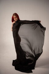 Naked girl with red hair wrapped in a black translucent transparent fabric fluttering in the wind, in the studio on a white background
