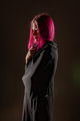 A girl with pink-crimson hair on a black background and darkness wrapped herself in a black cloth. Redemption, emotions, dark soul, religious cult