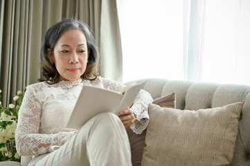 Beautiful Asian middle-aged woman sits on the comfortable sofa, reading a book.