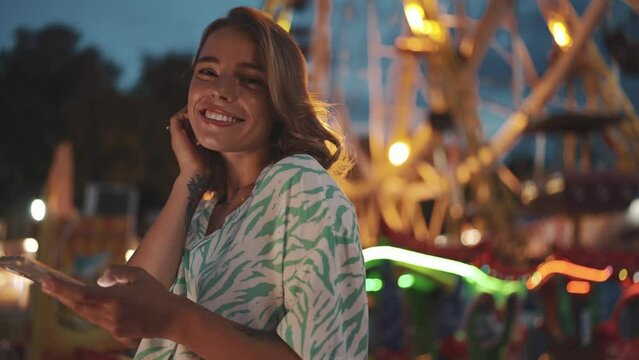 Positive young blonde woman typing on mobile and looking at the camera in adventure park at night