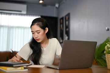 Fototapeta na wymiar Smart young Asian female college student concentrating doing her homework in living room