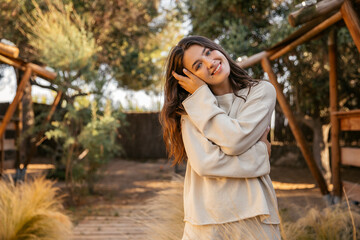 Tender young caucasian lady looks at camera, relaxes alone in fresh air. Girl with wavy hair wears casual clothes. Leisure lifestyle and beauty concept.