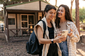 Cute caucasian young ladies are browsing social networks on phone relaxing in nature. Brunettes wear casual spring clothes. Technology, gadgets concept