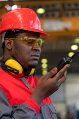 African American Worker In Protective Workwear Talking On Radio Communication Equipment In A Factory. Portrait Of Black Industrial Worker In Red Helmet, Safety Goggles And Safety Earmuffs.