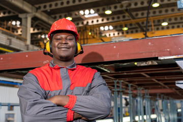 Happy African American Worker In Personal Protective Equipment Looking At Camera. Portrait Of Black Industrial Worker In Red Helmet, Hearing Protection Equipment And Work Uniform In A Factory.