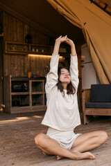 Fototapeta premium Slim young caucasian girl, closing eyes, does exercises, stretching arms up, sitting on floor. Brunette wears homemade casual clothes. Health concept